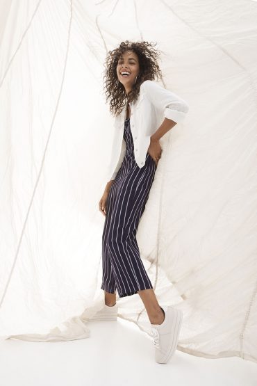 Smiling African-American woman in striped jumpsuit and white jacket posing in a fashion studio with white canvas backdrop and sneakers.