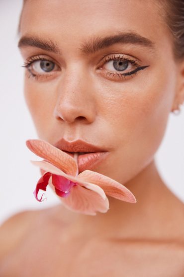 Close-up portrait of a woman with an orchid flower in her mouth against a white background, showcasing makeup details and natural light.