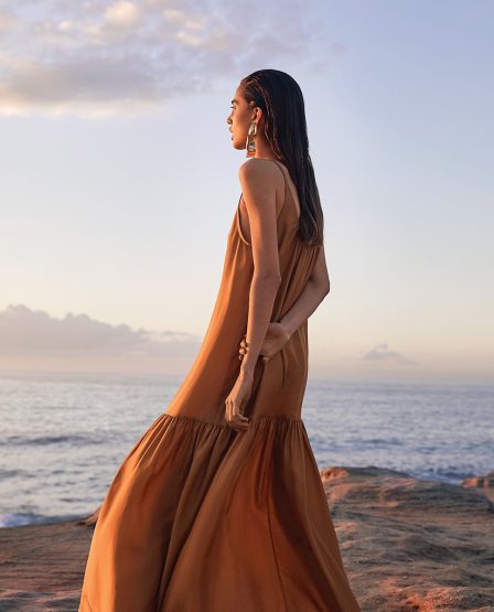 A woman in a flowing brown dress stands on a rocky outcrop overlooking the ocean at sunset, accessorized with large hoop earrings.