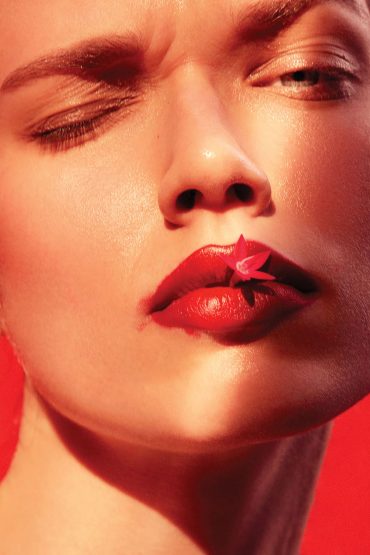 Close-up portrait of a woman with red lipstick and a small red flower held between her lips against a bold red background.