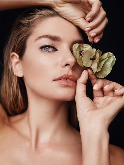 Close-up beauty portrait of a blonde woman with wet hair, makeup, and a plant near her face against a dark background.