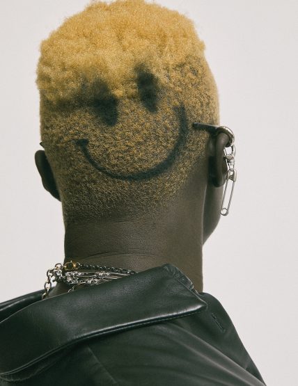 Close-up of a person's head and shoulders with a shaved, dyed-blonde hairstyle featuring a black smiley face design on the back of the head.