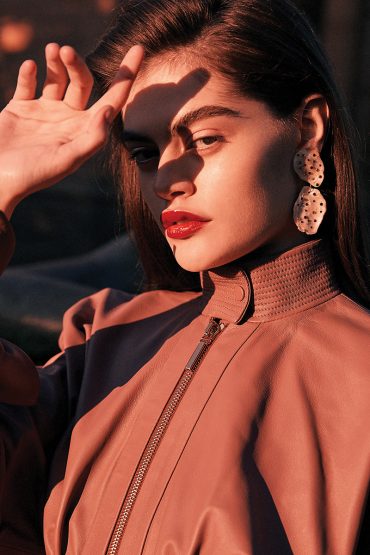 Portrait of a beautiful woman wearing statement earrings and a mauve zip-up jacket, hand raised to shield her face from the sunlight.