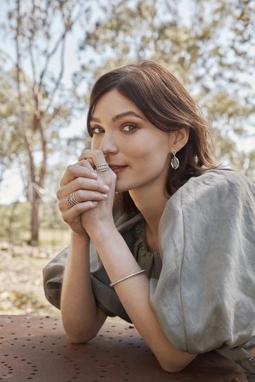Woman with dark hair and jewelry poses with hands clasped. She wears a light olive top in an outdoor setting with blurry trees.