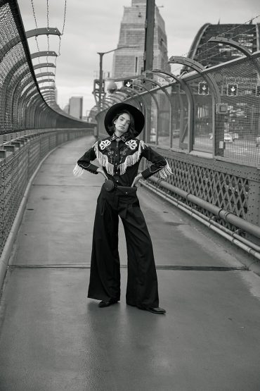Monochrome shot of a stylish woman in Western-inspired attire posing on a bridge with city buildings in the background on an overcast day.