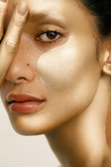 Close-up portrait of an adult female with golden skin, glancing at the camera, with a hand partially covering her face.