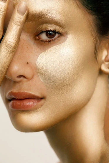 Close-up portrait of an adult female with golden skin, glancing at the camera, with a hand partially covering her face.