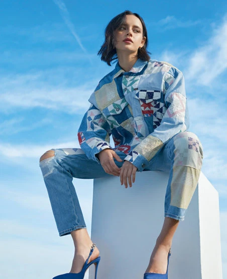A young woman in a patchwork denim jacket and jeans poses against a bright blue sky while seated on a white cube wearing blue pumps.