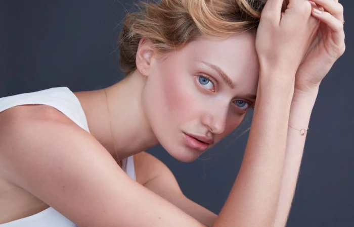 Close-up portrait of a woman with blue eyes wearing a white top and delicate jewelry against a dark grey background.