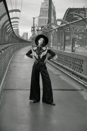 Model wearing a wide-brimmed hat, fringed western shirt, and wide-leg trousers standing on a bridge in Sydney.
