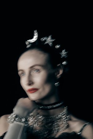 Elegant woman in a black top and ornate jewelry, wearing a celestial-themed headpiece, poses against a dark background with soft, diffused lighting.