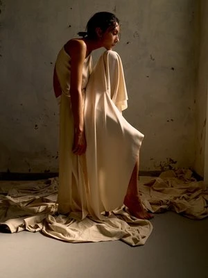Model wearing cream silk dress seated on draped fabric in a rustic room