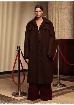 A woman in a brown coat stands indoors near red rope barriers, with a neutral background of striped walls and marble columns.