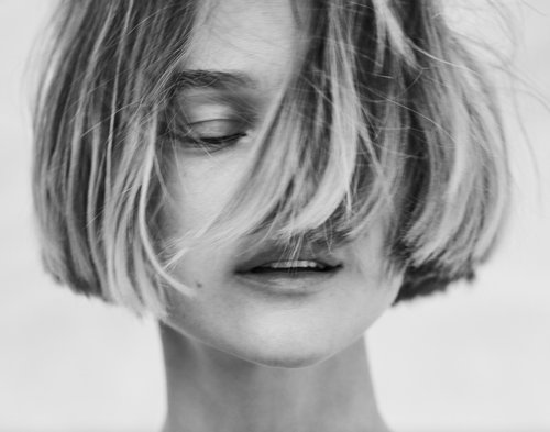 Close-up portrait of an adult woman with a blonde bob haircut, eyes closed, and diffused lighting, set against a blurred background.