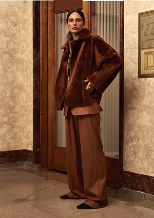 A woman poses in a brown fur coat and wide-legged trousers against a marble wall with wooden doors and framed art, fashion-forward style.