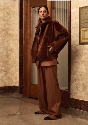 A woman poses in a brown fur coat and wide-legged trousers against a marble wall with wooden doors and framed art, fashion-forward style.