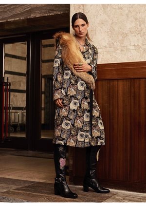 Woman in cat-print coat with fur stole and black boots stands in an indoor location with paneled walls and door frames.