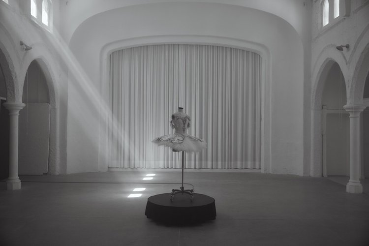 Ethereal monochrome shot of ballet tutu on stand in grand hall bathed in sunlight; white walls, archways, soft shadows.