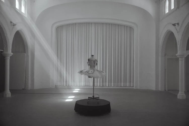 Ethereal monochrome shot of ballet tutu on stand in grand hall bathed in sunlight; white walls, archways, soft shadows.