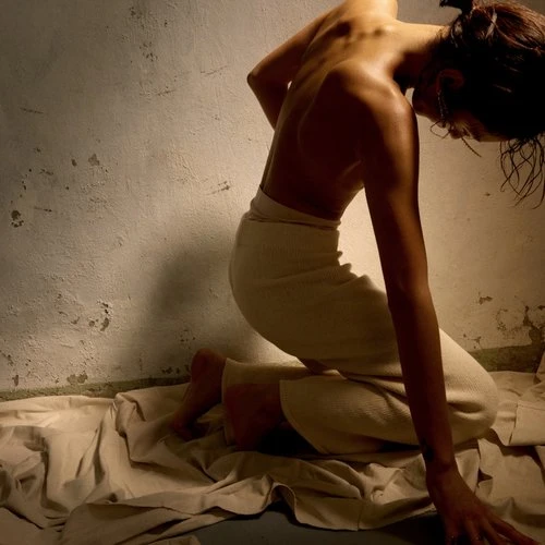 Back view of kneeling adult female with bare back, wearing light-colored pants, in front of a textured wall indoors on a sheet.