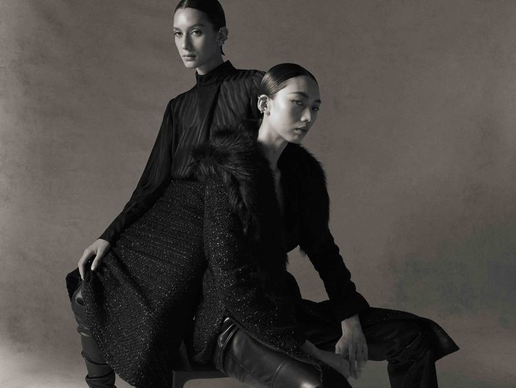 Two Asian women in dark, elegant fashion, one seated with a fur-trimmed coat, the other standing behind in a dark top, studio shot.