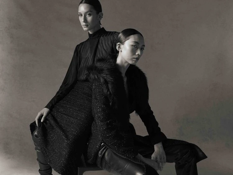 Two Asian women in dark, elegant fashion, one seated with a fur-trimmed coat, the other standing behind in a dark top, studio shot.