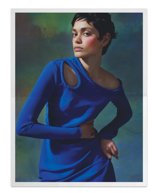 Portrait of woman with short dark hair in blue dress against green and blue mottled background, hands on waist looking at camera.