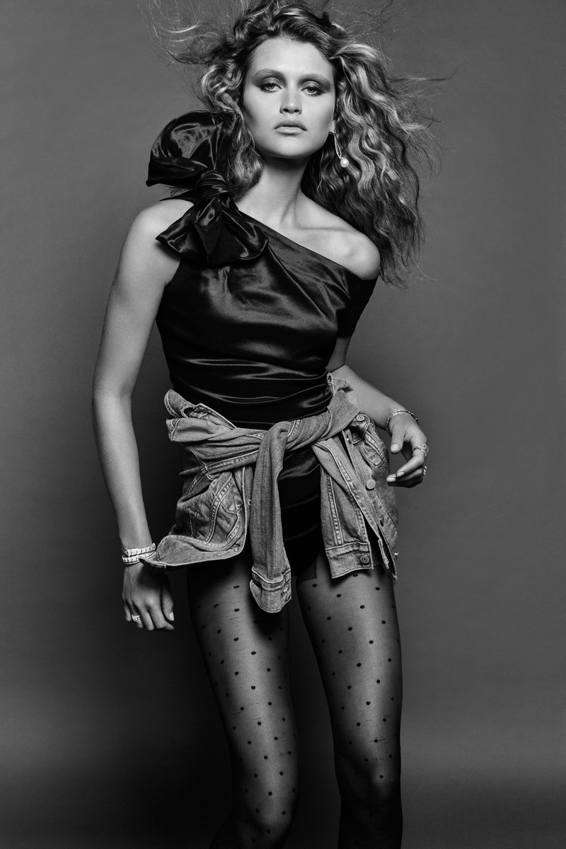 Black-and-white studio shot of a fashion model in a black top, denim jacket tied at the waist, and polka dot tights against a grey background.