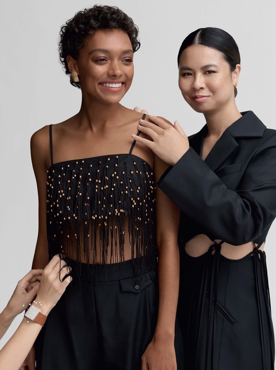 Two women pose indoors against a neutral backdrop. One wears a black, fringed top, while another wears a structured blazer with cutouts.
