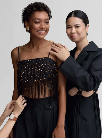 Two women pose indoors against a neutral backdrop. One wears a black, fringed top, while another wears a structured blazer with cutouts.