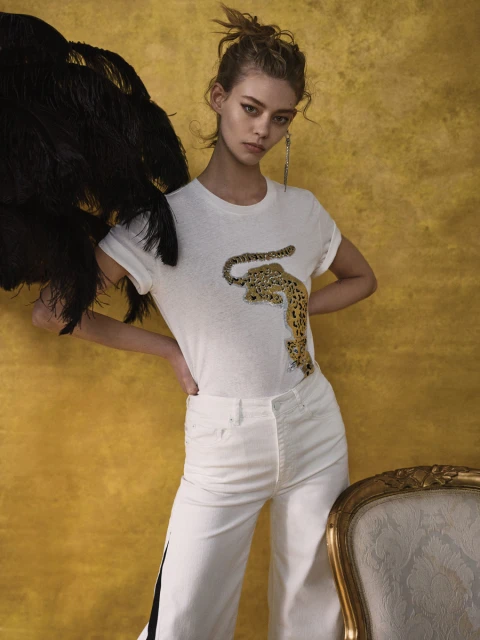 Fashionable young woman poses in white t-shirt with leopard graphic, wide leg pants, against a yellow wall and decorative chair.