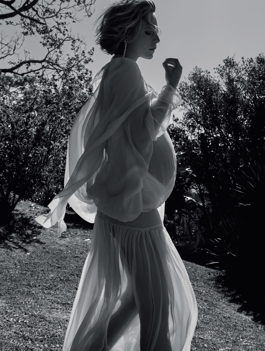 A pregnant woman in a flowing white dress stands in a grassy field, backlit by the sun, creating a serene and ethereal monochrome portrait.