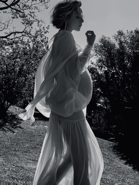 A pregnant woman in a flowing white dress stands in a grassy field, backlit by the sun, creating a serene and ethereal monochrome portrait.