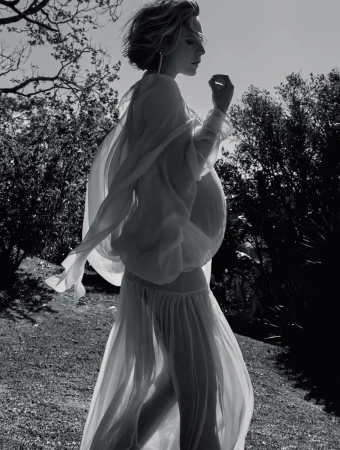 A pregnant woman in a flowing white dress stands in a grassy field, backlit by the sun, creating a serene and ethereal monochrome portrait.