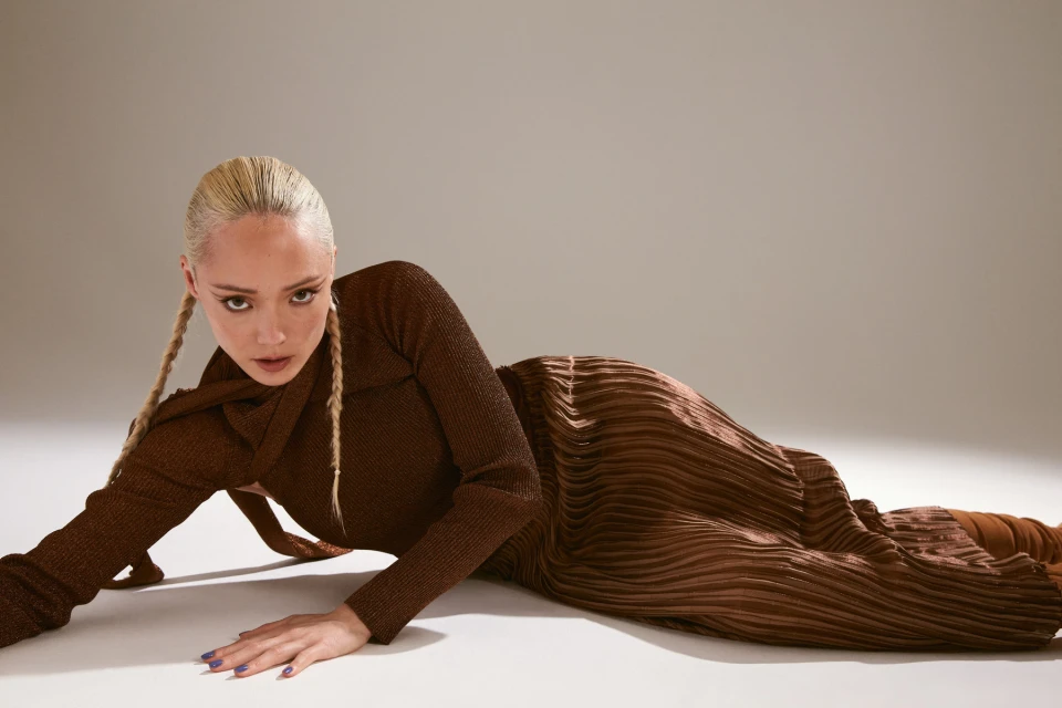 Woman with braided blonde hair poses in a long-sleeved brown pleated dress against a neutral backdrop, capturing fashion and style.
