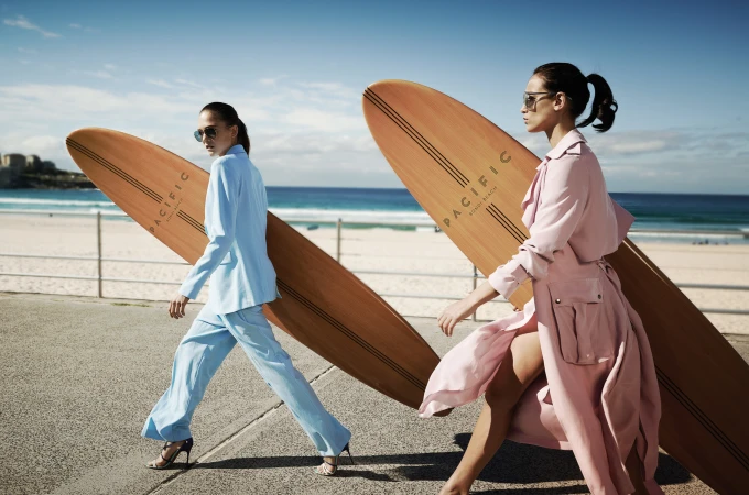 Two women in stylish coats walk with surfboards on a sunny beachside promenade, featuring ocean views and blue skies.