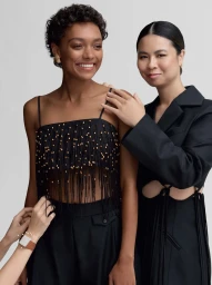Two women posing in black outfits against a white backdrop with a stylist adjusting clothing
