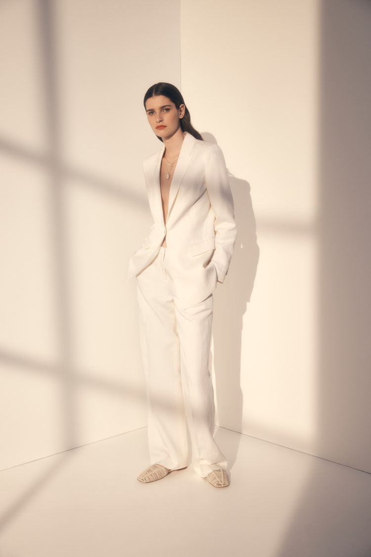 A woman in a white suit stands in a corner with soft, diffused light casting shadows, creating a minimalist and fashionable scene.