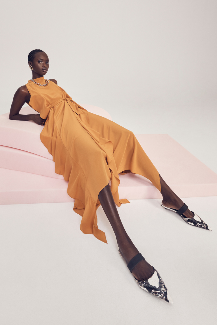 A stylish Black woman poses in a flowing orange dress and snake-print mules on a light pink surface, set against a white backdrop.