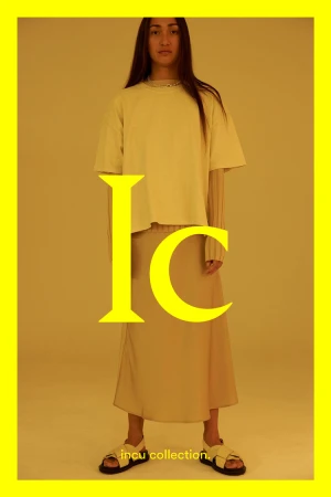 Model standing in cream t-shirt, long skirt, and sandals against a warm yellow gradient background with bold yellow text overlay.