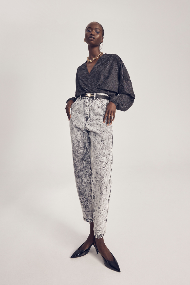 An adult woman with short hair stands with hands on hips, modeling high-waisted jeans, a metallic top, and high-heeled shoes in a studio.