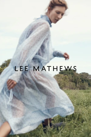 A woman in a sheer light blue patterned dress walking through a grassy field under an overcast sky.