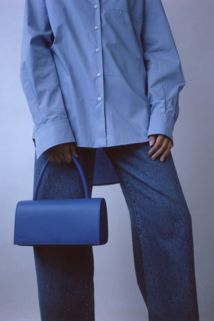 Woman in blue oversized shirt and wide-leg jeans holds blue bag in studio setting.