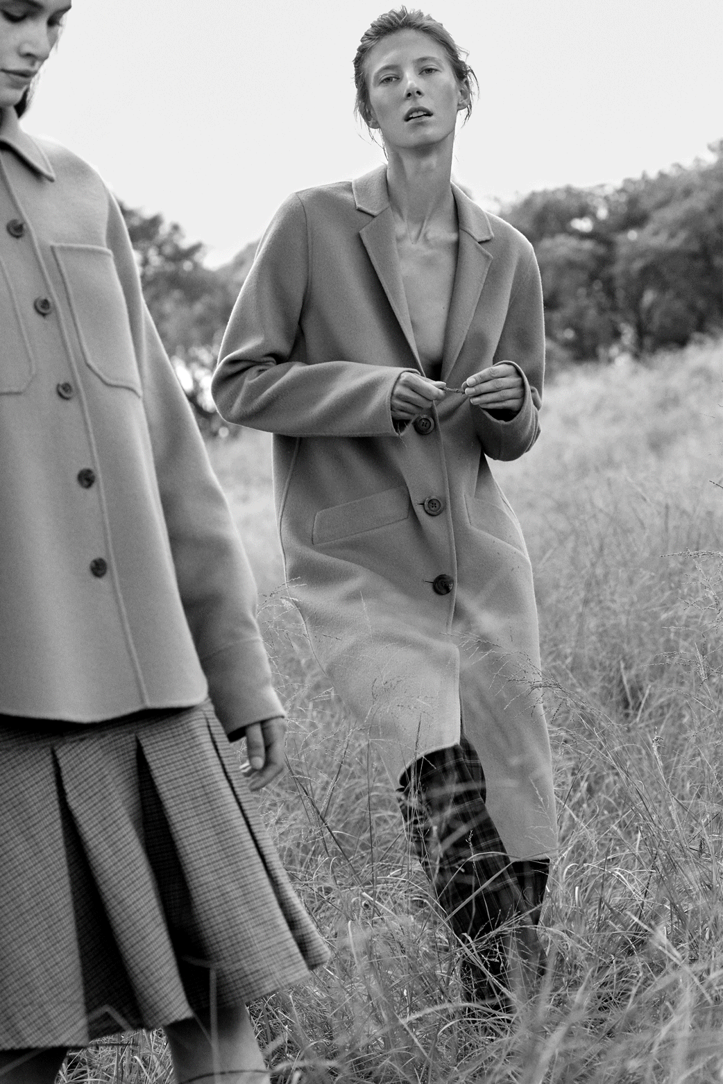 Two women in fashionable dresses pose in a grassy field on a sunny day. One wears a pink jacket, the other a ruffled dress.