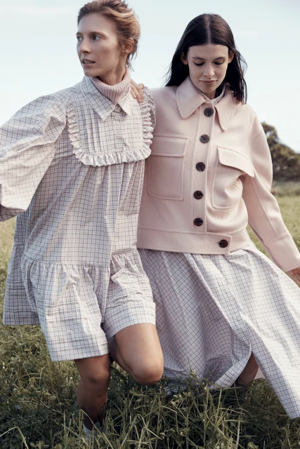 Two women in fashionable dresses pose in a grassy field on a sunny day. One wears a pink jacket, the other a ruffled dress.