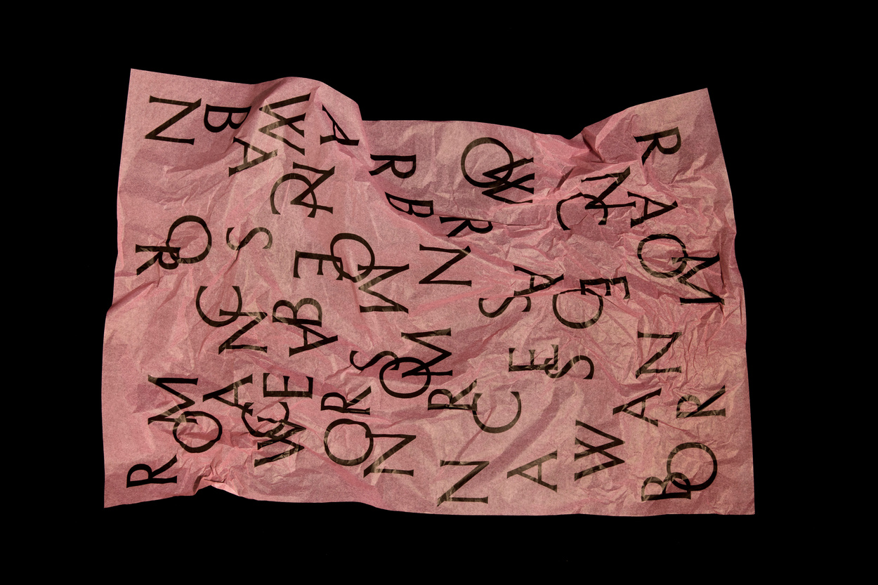 Wrinkled light pink paper with overlapping black letters on a dark background, creating an abstract graphic design texture.