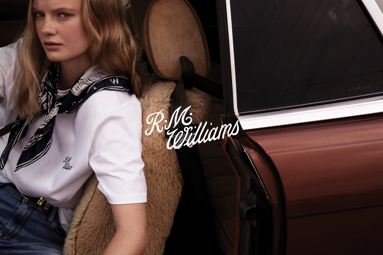 Portrait of blonde woman sitting inside classic car wearing a white tee with logo, scarf, and jeans. The brand logo R.M. Williams is visible.