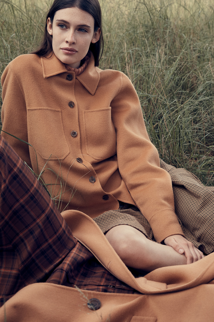 A fair-skinned woman with long dark hair sits in a field of tall grass wearing an orange jacket and plaid blanket in natural light.