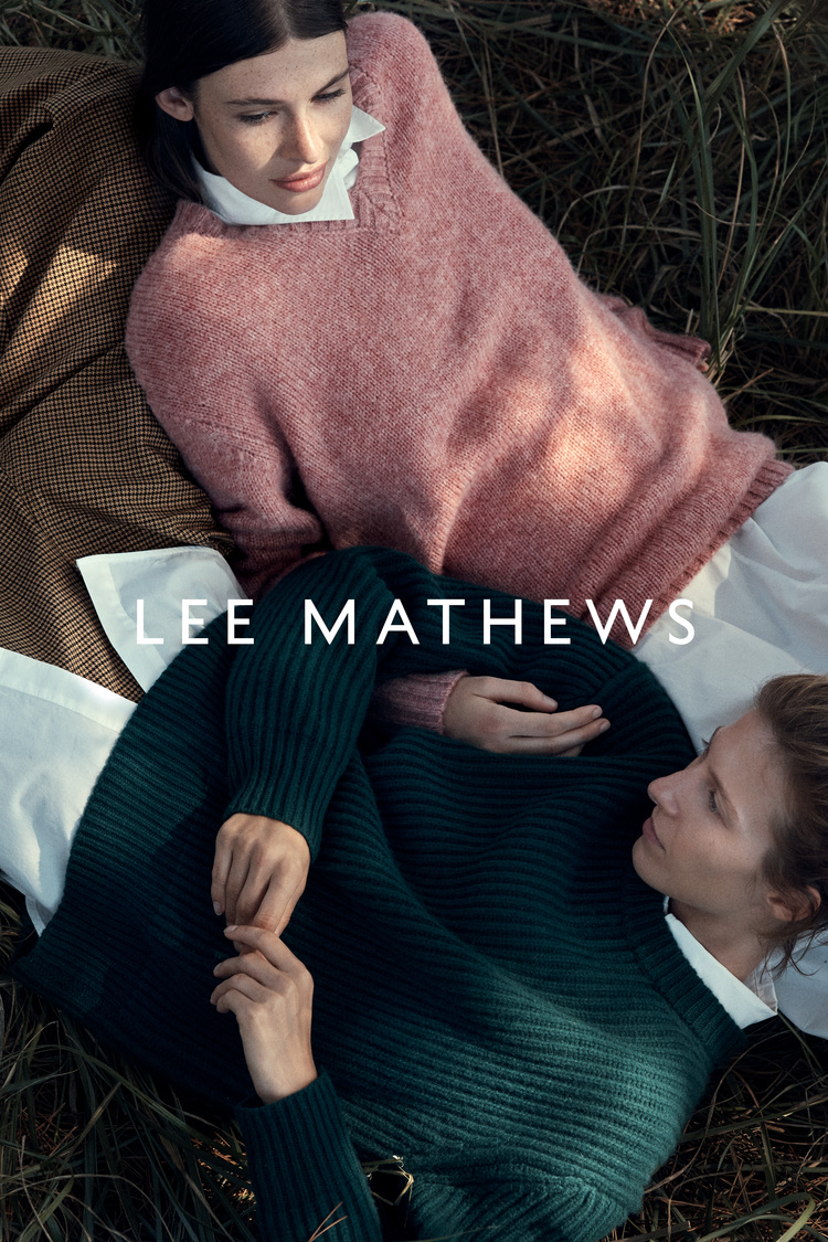 Two women lying in tall dry grass wearing knit sweaters and white button-down shirts outdoors