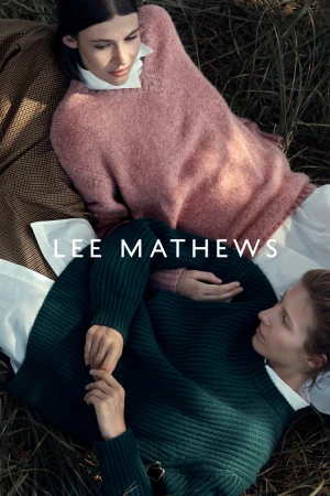 Two women lying in tall dry grass wearing knit sweaters and white button-down shirts outdoors